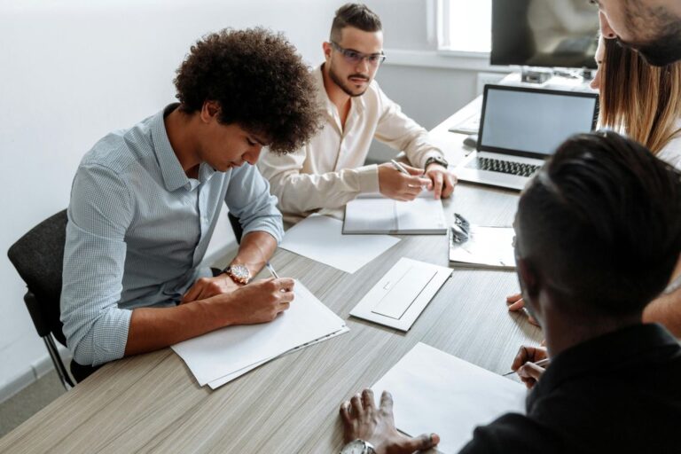Equipe de quatro profissionais em reunião de trabalho em escritório de PME, discutindo processo comercial com documentos e laptop