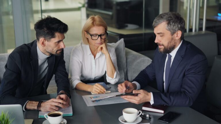 Dois executivos e uma executiva em reunião de negócios analisando relatório de vendas sobre mesa em escritório moderno com divisórias de vidro