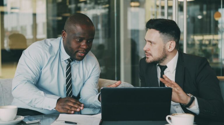 Dois executivos colaboram em laptop discutindo relatório em sala de reuniões com parede de vidro