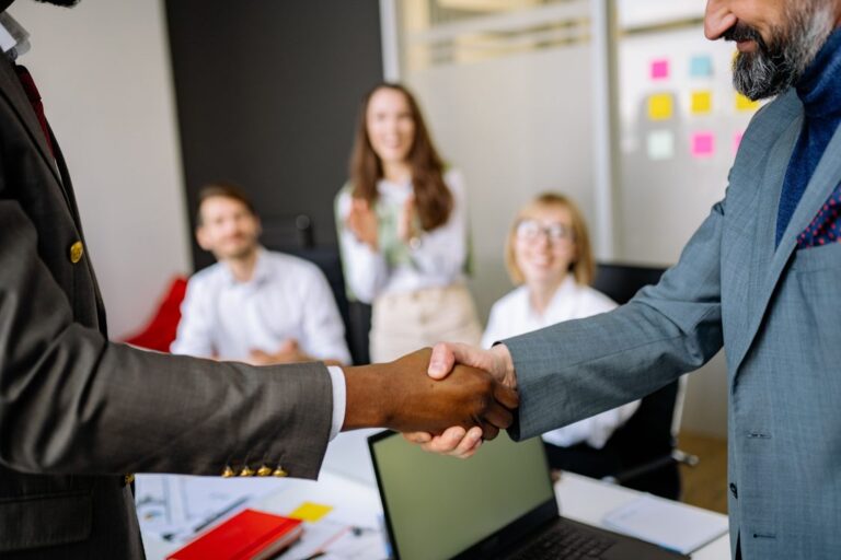 Dois executivos B2B realizando handshake em sala de reunião enquanto equipe diversa aplaude ao fundo, laptop e documentos sobre a mesa
