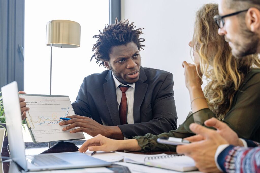 Profissional de vendas negro de terno apresentando gráfico e gesticulando com caneta durante pitch para dois clientes sentados à mesa em reunião de negócios no escritório