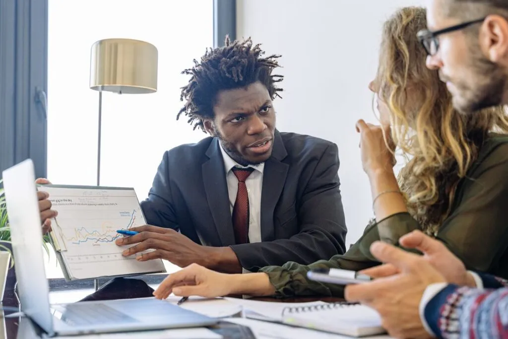 Profissional de vendas negro de terno apresentando gráfico e gesticulando com caneta durante pitch para dois clientes sentados à mesa em reunião de negócios no escritório