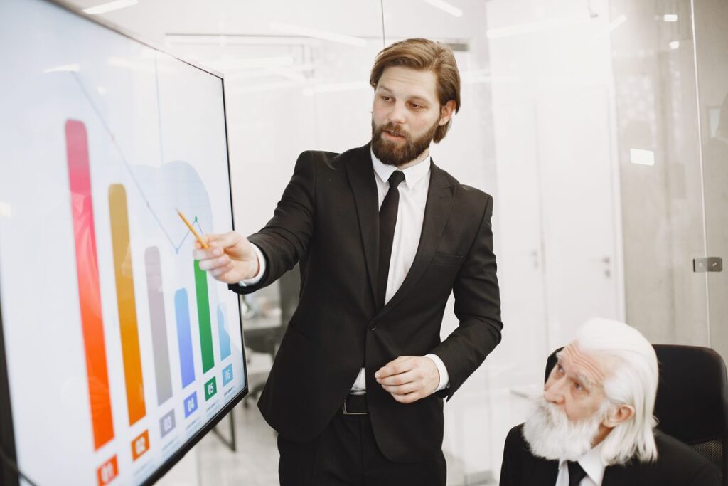 Vendedor de terno apontando dados de crescimento em tela para executivo sênior durante apresentação de proposta comercial B2B