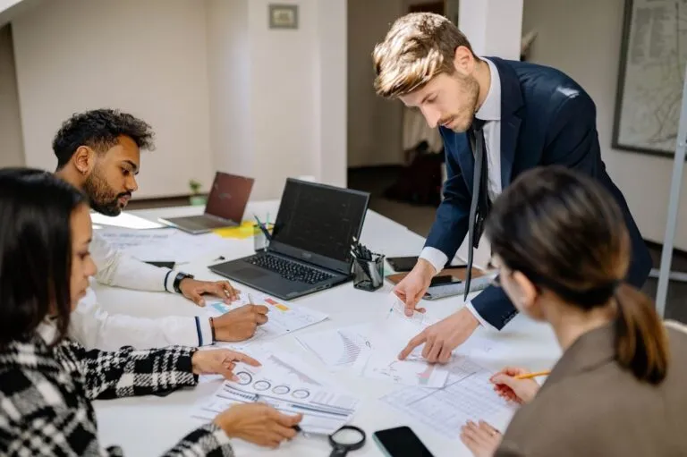 Equipe revisando relatórios impressos com gráficos ao redor de uma mesa, com o gestor em pé apontando um dos papéis