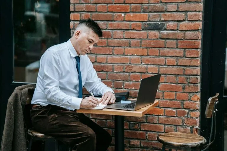 Profissional de camisa e gravata escrevendo em um caderno ao lado do notebook, numa mesa de café